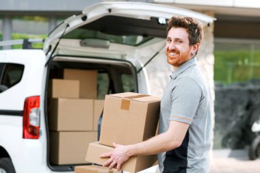 Kutuları taşıyan gülümseyen profesyonel teslimatçı, paketleri bir minivana yüklüyor.