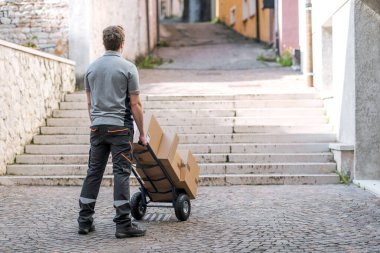 Teslimatçı, şehir caddesinde kutularla dolu bir el arabasını itiyor. Yolda merdivenleri buluyor ve duruyor.