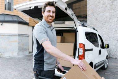 Şehirde paket teslimatı yapan gülümseyen bir kuryenin portresi, paketlerle dolu bir el kamyonu itiyor.