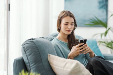 Evdeki kanepede oturan ve akıllı telefonuyla iletişim kuran mutlu genç kadın sohbet ediyor ve mobil uygulamalar kullanıyor.