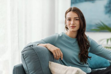 Oturma odasındaki kanepede oturan ve kameraya gülümseyen güzel genç bir kadın.