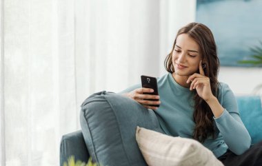 Evdeki kanepede oturan ve akıllı telefonuyla iletişim kuran mutlu genç kadın sohbet ediyor ve mobil uygulamalar kullanıyor.