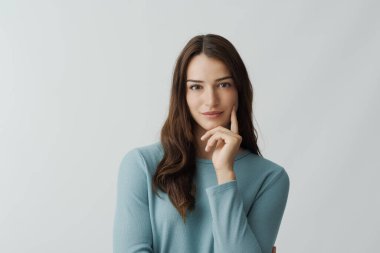 Çekici genç bir kadın, eli çenesinde poz veriyor ve beyaz arka planda izole bir şekilde kameraya bakıyor.