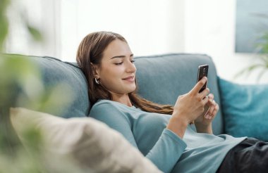 Mutlu genç bir kadın evdeki kanepede dinleniyor ve dokunmatik ekranlı akıllı telefonu, teknolojisi ve yaşam tarzı konseptiyle sohbet ediyor.