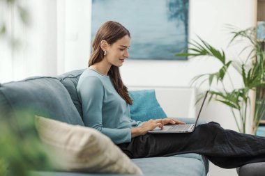 Güzel genç bir kadın oturma odasındaki kanepede dinleniyor ve dizüstü bilgisayarını, yaşam tarzını ve teknoloji konseptini kullanıyor.