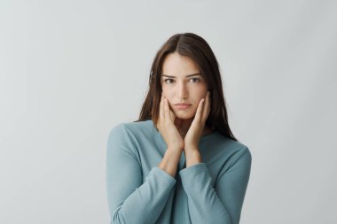 Genç bir kadın yüzüne dokunuyor ve beyaz arka planda izole bir şekilde kameraya bakıyor.