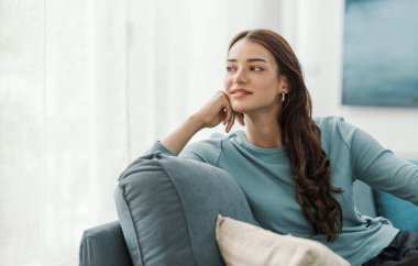 Güzel bir genç kadının portresi kanepede oturuyor ve evde dinleniyor, başka yere bakıyor ve gülümsüyor.