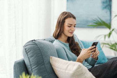 Evdeki kanepede oturan ve akıllı telefonuyla iletişim kuran mutlu genç kadın sohbet ediyor ve mobil uygulamalar kullanıyor.