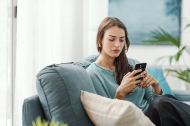Evdeki kanepede oturan ve akıllı telefonuyla iletişim kuran mutlu genç kadın sohbet ediyor ve mobil uygulamalar kullanıyor.
