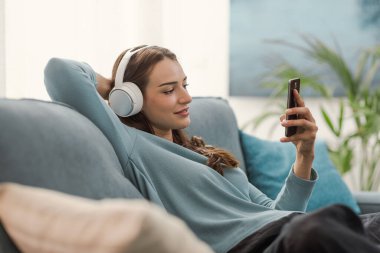 Evdeki kanepede oturan mutlu genç kadın, kulaklık takıyor ve akıllı telefonu, eğlence ve teknoloji konseptiyle video oynuyor.