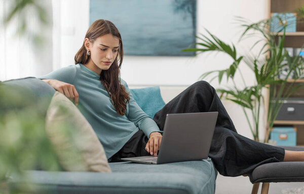 Красивая молодая женщина отдыхает на диване в гостиной и с помощью ноутбука, образа жизни и технологии