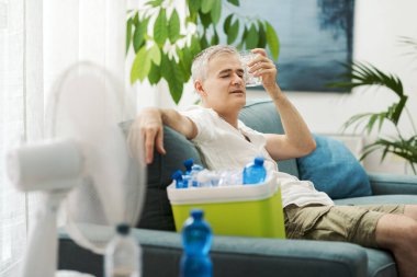 Evdeki kanepede su şişeleri ve elektrikli vantilatörle dolu bir soğutucunun yanında oturan bitkin adam sıcaktan muzdarip.