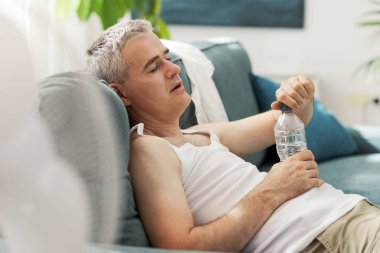 Yorgun bir adam sıcak hava dalgası sırasında evdeki kanepede dinleniyor, su içiyor ve sıcaktan acı çekiyor.