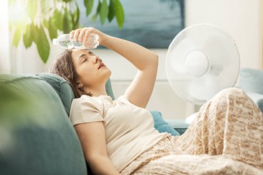 Young woman sitting on the couch at home and keeping a fresh water bottle on her forehead, she is suffering from the heat clipart