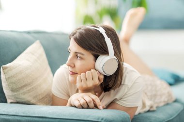 Güzel sakin bir kadın kanepede uzanıyor ve kulaklık takıyor, müzik dinliyor ve başka tarafa bakıyor.