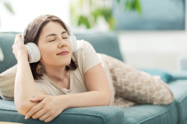 Güzel, sakin bir kadın kanepede uzanıyor ve kulaklık takıyor. Gözleri kapalı müzik dinliyor.