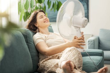 Evdeki sıcaktan muzdarip bir kadın, koltukta oturuyor ve yüzüne elektrikli vantilatör kullanarak temiz hava üflüyor.