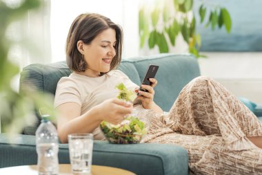 Kanepede oturup öğle yemeği arası veren mutlu kadın salata yiyor ve akıllı telefonunu kullanıyor.