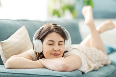 Güzel, sakin bir kadın kanepede uzanıyor ve kulaklık takıyor. Gözleri kapalı müzik dinliyor.