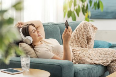 Mutlu kadın evdeki kanepesinde dinleniyor, kulaklık takıyor ve akıllı telefonunu kullanıyor.
