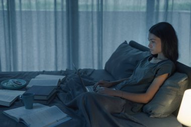 Yatakta oturan ve dizüstü bilgisayarında yazan kız öğrenci, bütün gece çalışıyor ve sınavdan önce ders çalışıyor.