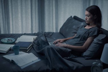 Yatakta oturan ve dizüstü bilgisayarında yazan kız öğrenci, bütün gece çalışıyor ve sınavdan önce ders çalışıyor.