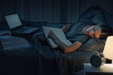 Exhausted female student falling asleep while studying a textbook in bed, she is preparing for the test clipart