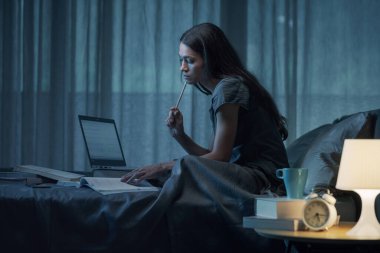 Odaklanmış öğrenci gece geç saatlere kadar çalışıyor, yatakta oturuyor ve ders kitabı okuyor.