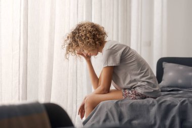 Mutsuz bir kadın uyanıyor ve yatağında oturuyor. Kafasına dokunuyor.