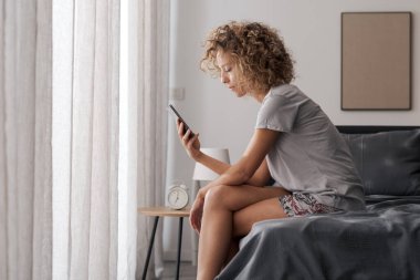 Genç ve çekici bir kadın sabah yatağında oturmuş akıllı telefonunu kontrol ediyor.