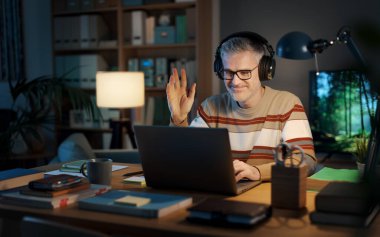 Evdeki masasında oturan bir adam dizüstü bilgisayarında, iletişim ve teknoloji konseptinde bir video görüşmesi yapıyor.