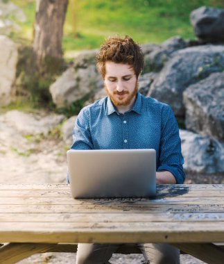 Genç hipster adam açık havada doğa, ağaçlar ve bitkiler üzerinde arka plan içinde onun laptop üzerinde çalışıyor