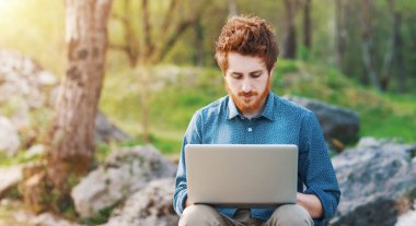 Genç hipster adam açık havada doğa, ağaçlar ve bitkiler üzerinde arka plan içinde onun laptop üzerinde çalışıyor