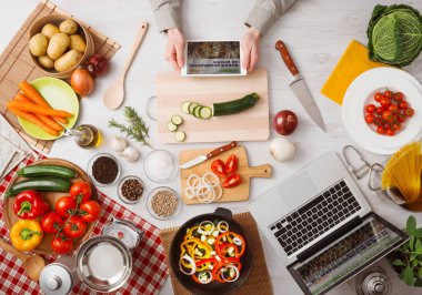 Mutfakta yemek yapan adam elinde gıda malzemeleri, mevsimlik sebzeler ve çatal bıçaklarla dijital tablet tutuyor.