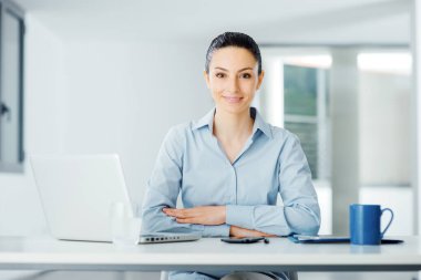 Güzel genç iş kadını ofis masasında oturuyor ve kameraya gülümsüyor.
