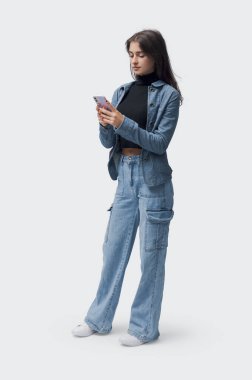 Genç ve şık bir kadın ayakta duruyor ve akıllı telefonunu kontrol ediyor, uzun boylu, beyaz arka planda soyutlanmış.