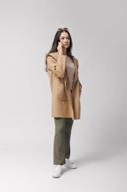 Genç ve şık bir kadın ayakta duruyor ve akıllı telefonuyla telefon görüşmesi yapıyor, uzun boylu, beyaz arka planda soyutlanmış.