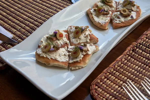 Zarif aperatif tostlar beyaz seramik bir tabakta dokunmuş bir servis altlığı üzerinde servis edilir. Her kızarmış parçanın üstünde krem peynir, yeşil zeytin, şifalı otlar ve yenilebilir çiçekler var.