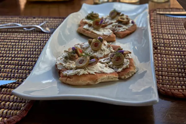 Zarif aperatif tostlar beyaz seramik bir tabakta dokunmuş bir servis altlığı üzerinde servis edilir. Her kızarmış parçanın üstünde krem peynir, yeşil zeytin, şifalı otlar ve yenilebilir çiçekler var.