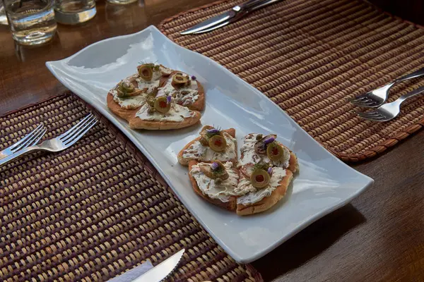 Zarif aperatif tostlar beyaz seramik bir tabakta dokunmuş bir servis altlığı üzerinde servis edilir. Her kızarmış parçanın üstünde krem peynir, yeşil zeytin, şifalı otlar ve yenilebilir çiçekler var.