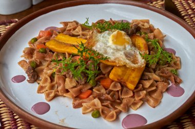 Yumuşak sığır eti, bezelye, havuç ve kızarmış muz ve kızarmış yumurtayla taçlandırılmış lezzetli bir sosla pişirilmiş papyon makarnası. Taze otlar ve mor soslarla süslenmiş..