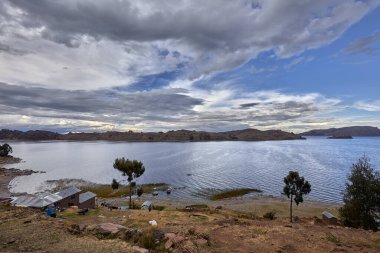 Capachica Yarımadası 'nın kuzeyindeki Titicacas Gölü manzarası Andean tepelerini, derin mavi suları ve dramatik fırtınalı gökyüzünün altında geleneksel tarım arazilerini gösteriyor..