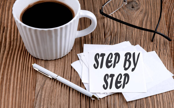 STEP BY STEP text on a sticky with coffee and pen on wooden background