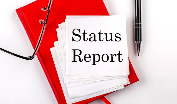 STATUS REPORT text on sticker on a red notebook with pen and glasses