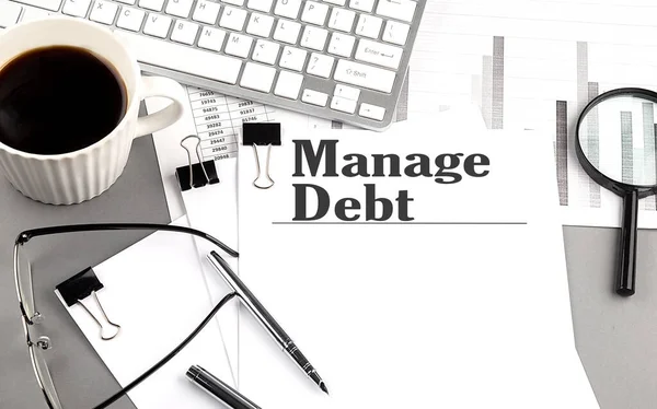 MANAGE DEBT text on a paper with magnifier, coffee and keyboard on a grey background