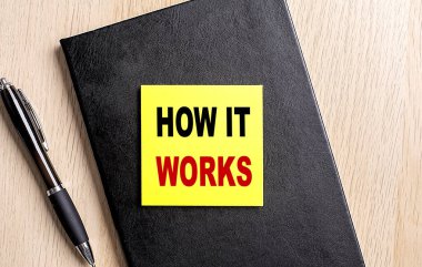 HOW IT WORKS text on sticky on black notebook with pen