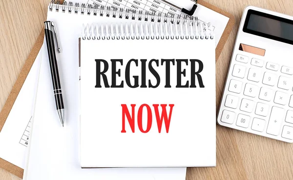 REGISTER NOW is written in white notepad near calculator, clipboard and pen. Business concept