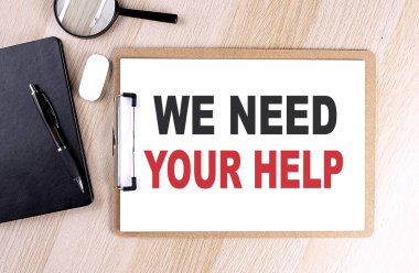 WE NEED YOUR HELP text on a clipboard on wooden background