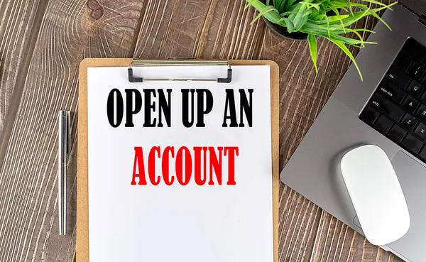 OPEN UP AN ACCOUNT text on a paper clipboard with laptop and mouse on wooden background , business concept