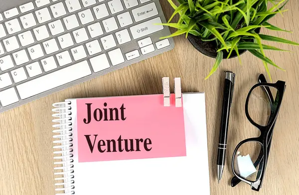 stock image JOINT VENTURE text pink sticky on a notebook with keyboard, pen and glasses . 